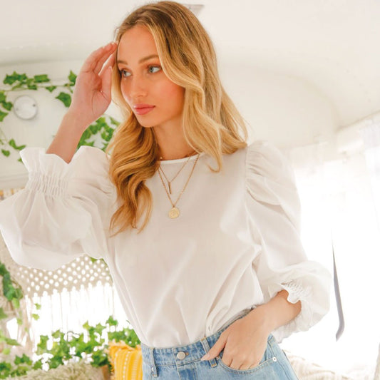 Woman wearing a white blouse with long sleeves and blue jeans, standing in a bright room with plants.